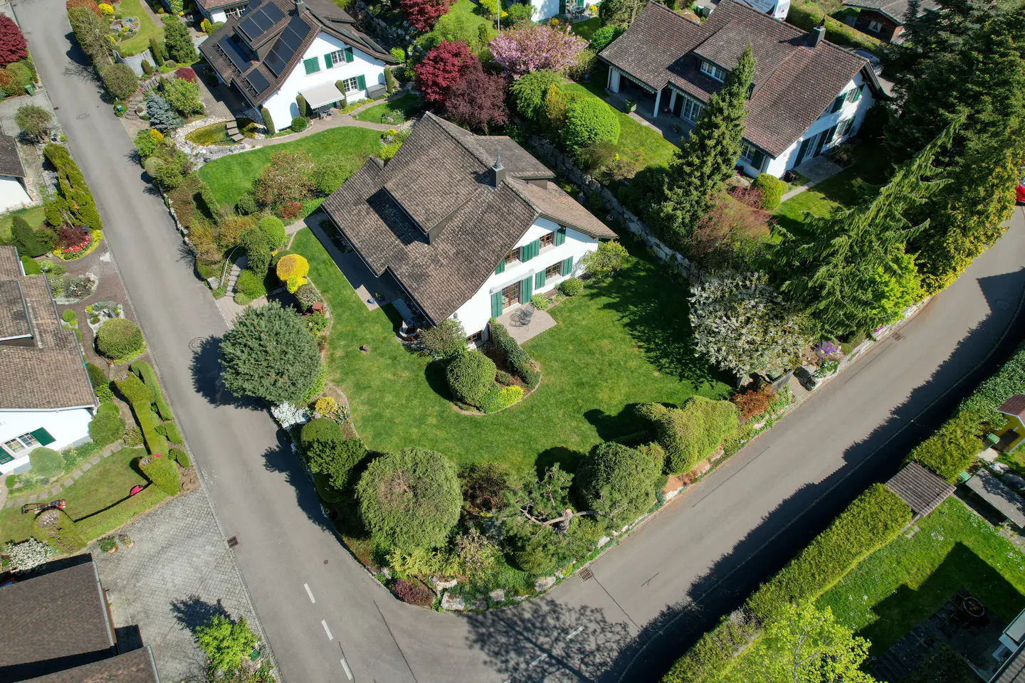 Aerial view of a white house with green shutters and a brown roof, surrounded by a lush green lawn and colorful trees.
