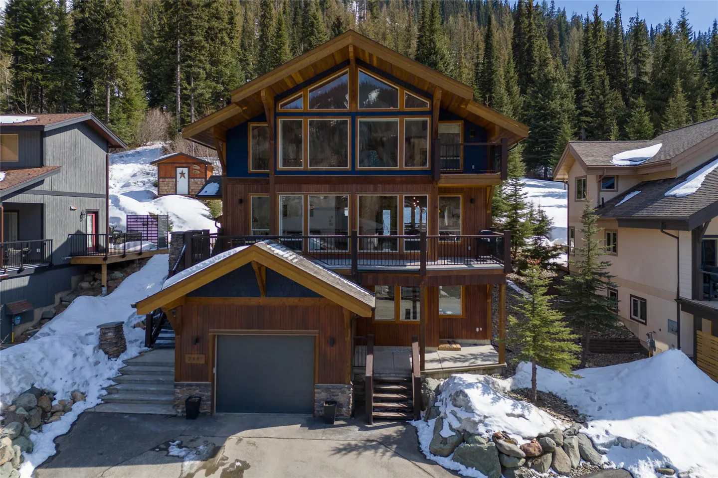 A three-story wood cabin with a garage and a balcony, surrounded by snow and trees.
