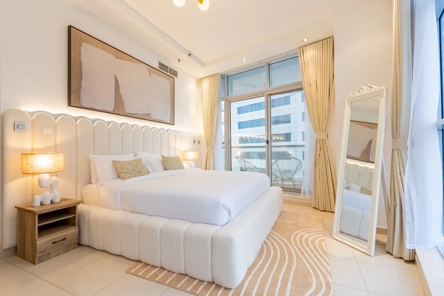 Bright bedroom with a white bed, headboard, and linens. A large window with curtains overlooks a city view. A full-length mirror stands near the window.