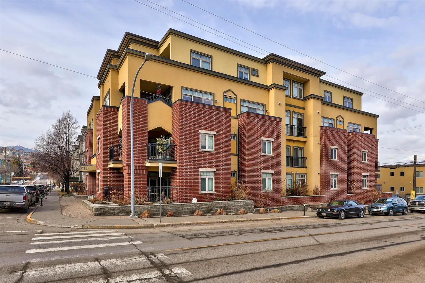 Street view of a yellow and brick apartment building with balconies, cars parked on the street, and a crosswalk in front.