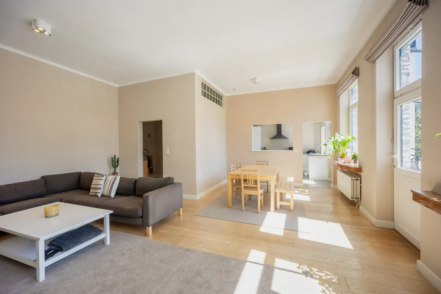 Bright, open-plan living space with a gray sectional sofa, white coffee table, and a wooden dining set. Large windows provide natural light.
