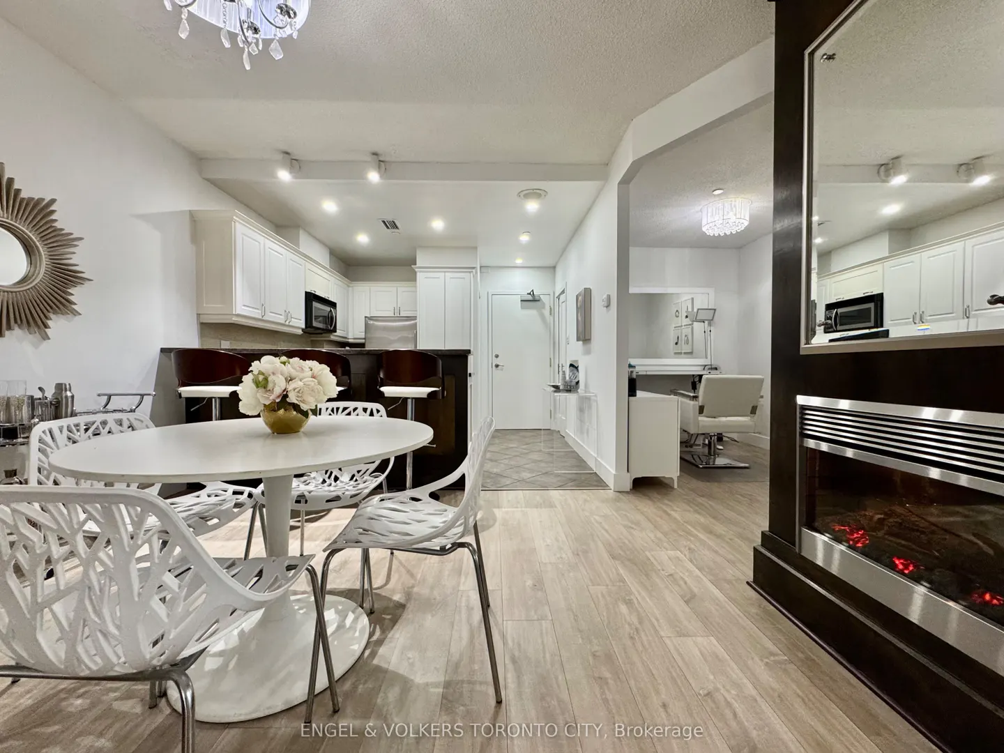Modern dining area with white tulip table, intricate white chairs, and a fireplace, leading to a kitchen and salon space.