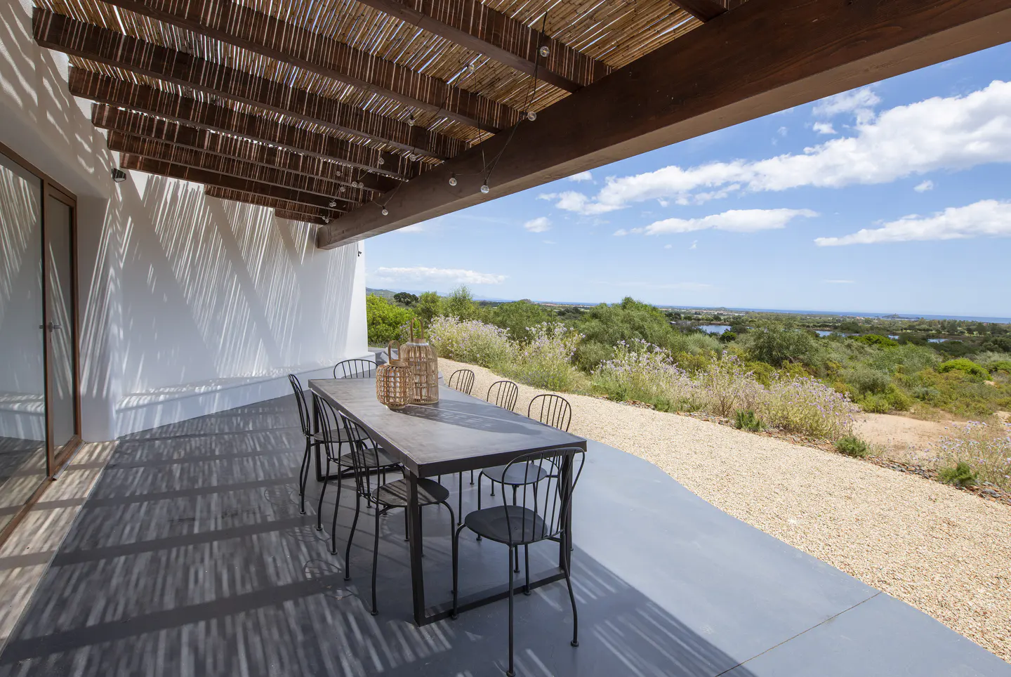 Outdoor patio with a long table and chairs under a thatched roof, overlooking a green landscape and blue sky.