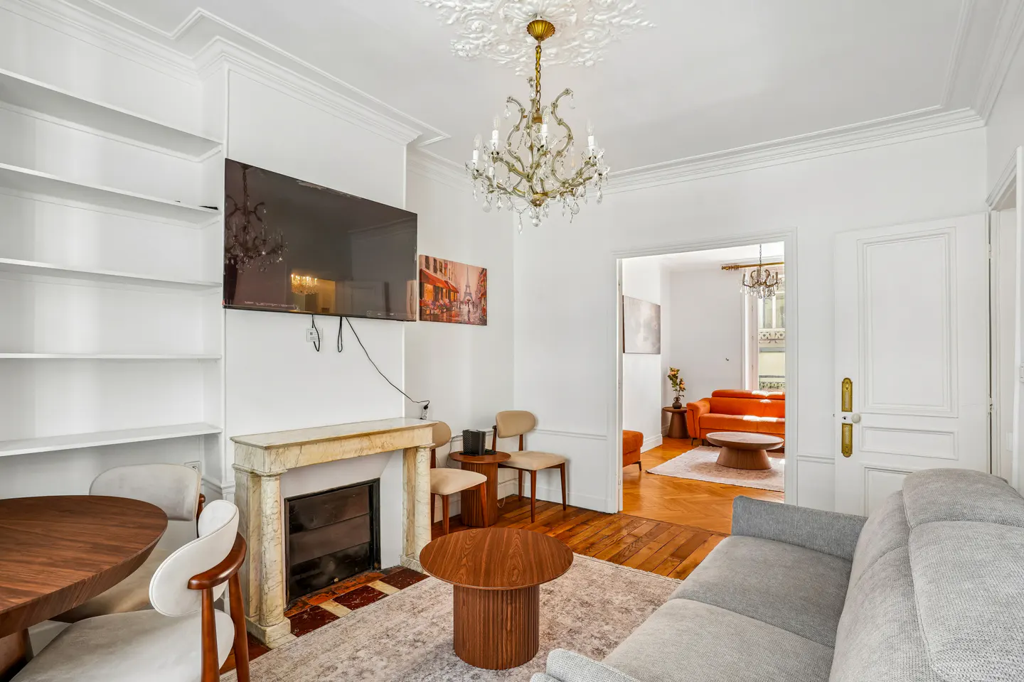 Bright living room with white walls, hardwood floors, and a gray sofa. A fireplace, TV, and chandelier add elegance. Another room is visible through a doorway.
