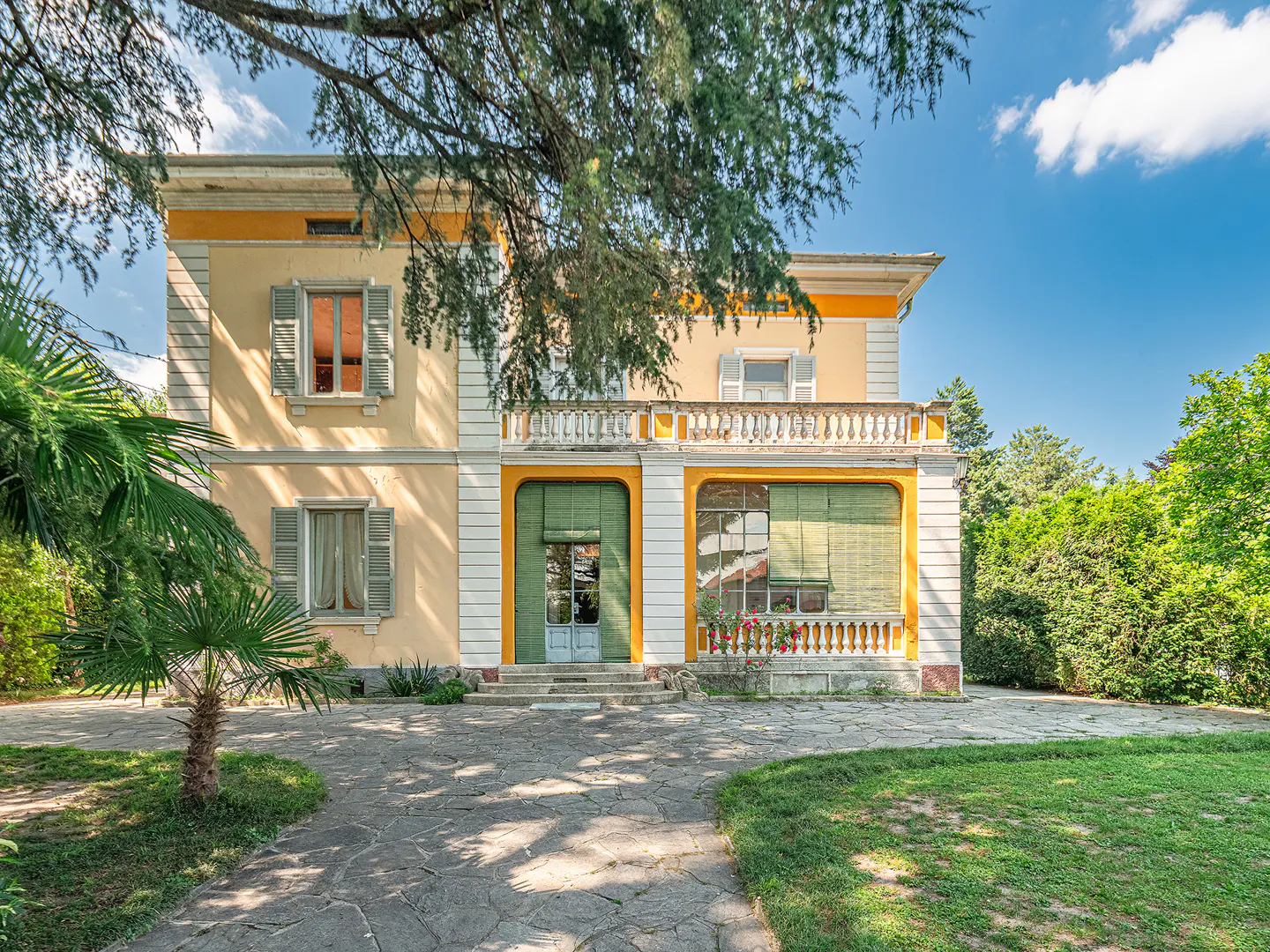 A two-story yellow villa with green shutters, a stone patio, and lush garden under a bright blue sky.