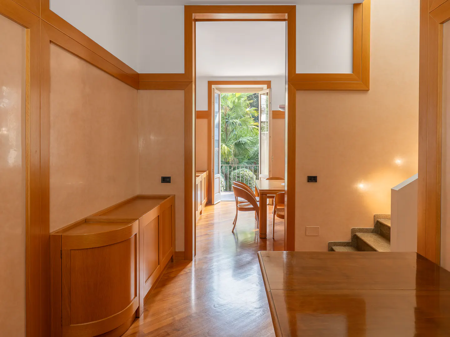Hallway with peach walls and wood trim leading to a dining room with a balcony view of lush green trees.