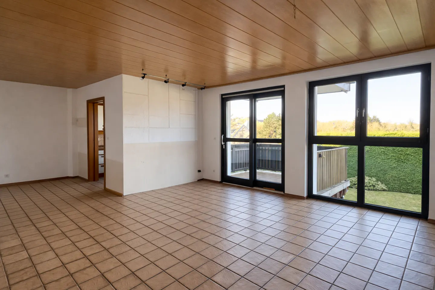 Empty room with tan tile floor, wood ceiling, and white walls. Black framed windows and doors lead to a balcony and garden.
