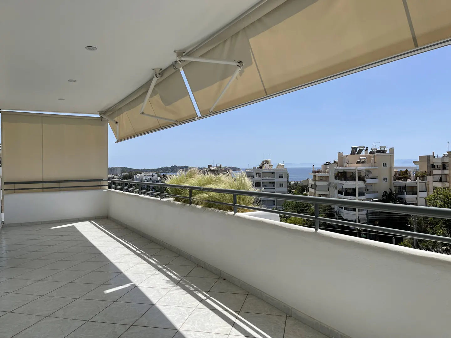 Balcony with beige awning, white railing, and tile floor. City buildings and blue sea are visible in the background.
