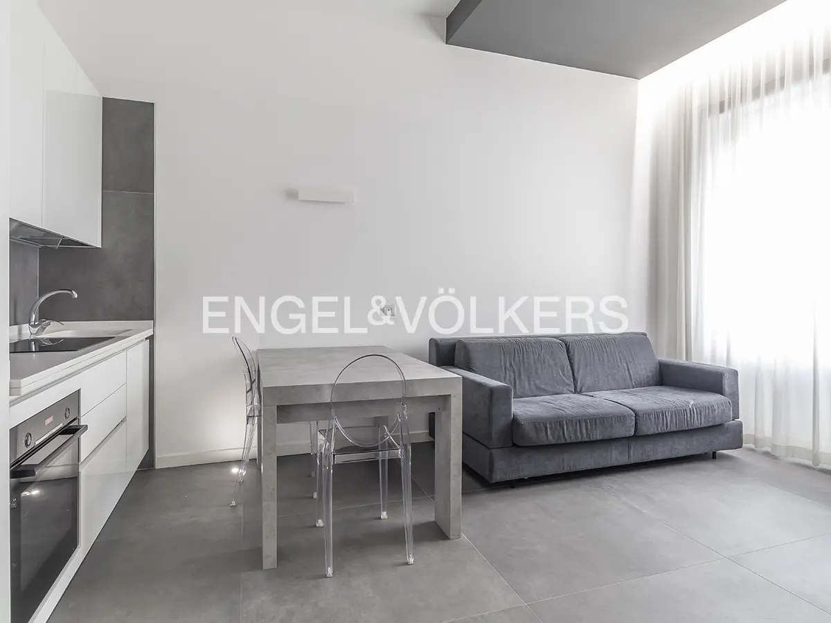 Modern studio apartment with a grey sofa, concrete dining table, clear chairs, and a sleek white kitchen with grey tiles.