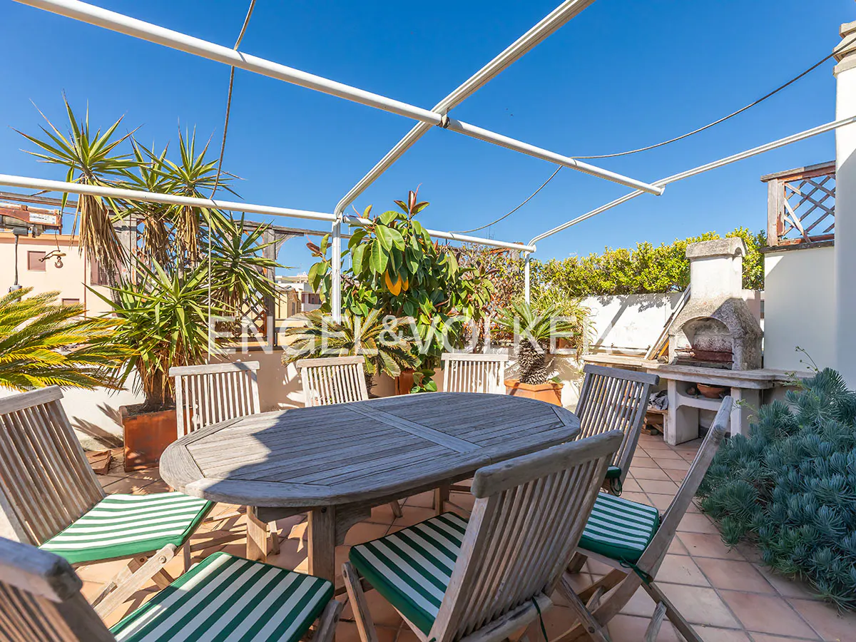 Outdoor patio with a wooden table, chairs with green and white striped cushions, and potted plants under a white pergola against a blue sky.