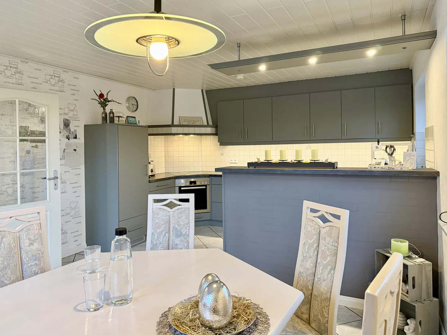 A kitchen and dining area with gray cabinets, white walls, and a white table with chairs. A modern light fixture hangs above the table.