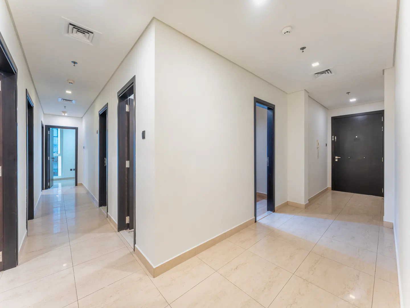 A long, bright hallway with beige tile floors and white walls. Dark brown doors line both sides of the hallway.