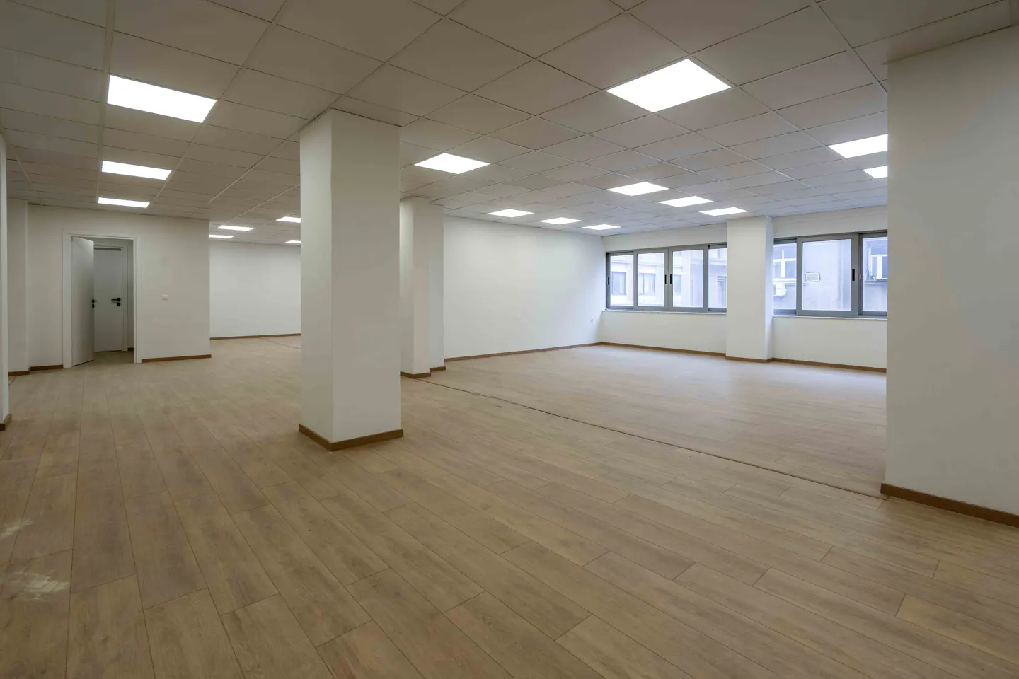 Bright, empty office space with wood floors, white walls, and square ceiling lights. Windows line one wall, and support columns are spaced throughout.
