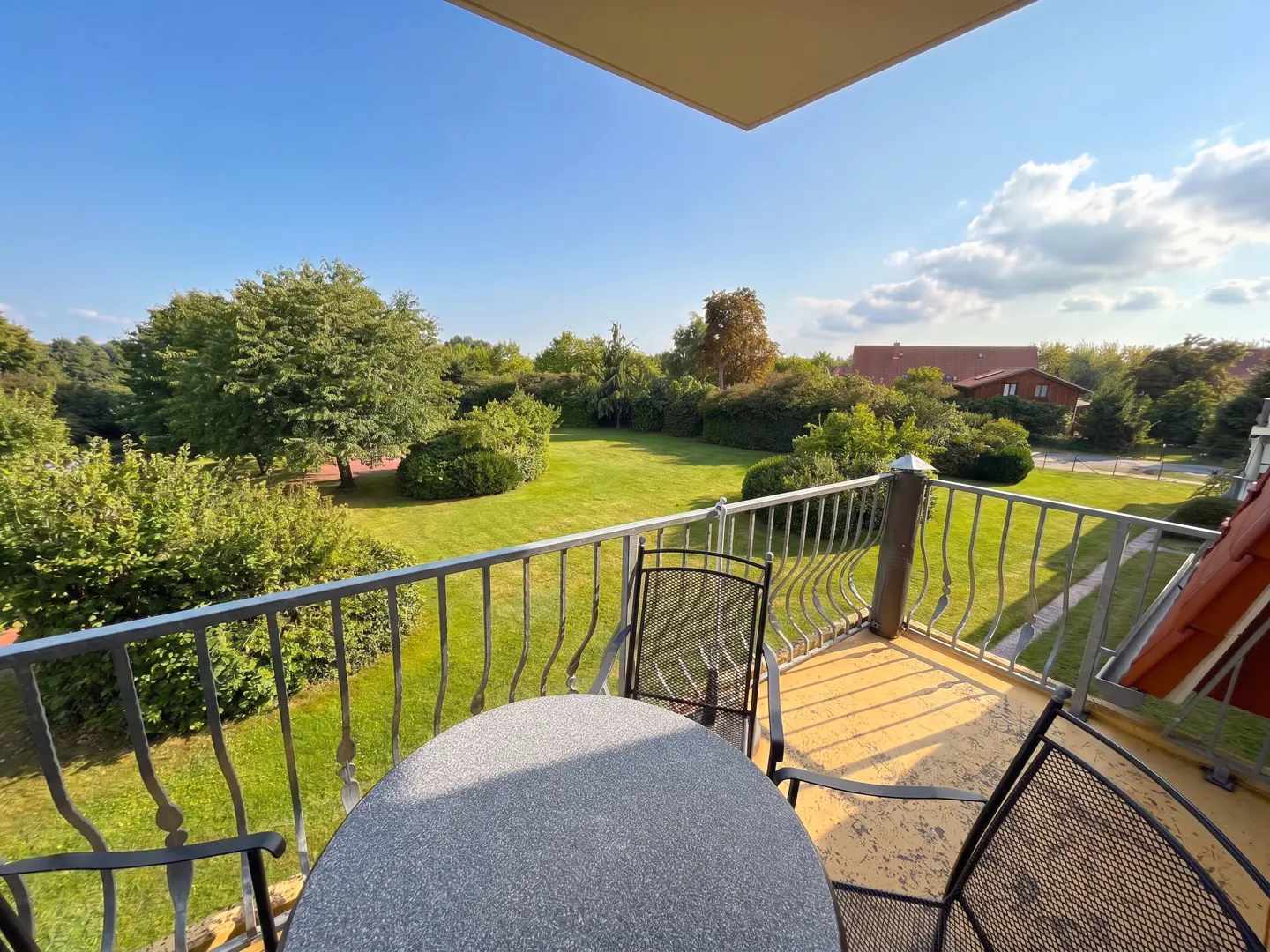 Balcony view with metal table and chairs overlooking a green lawn, trees, and a house under a blue sky.