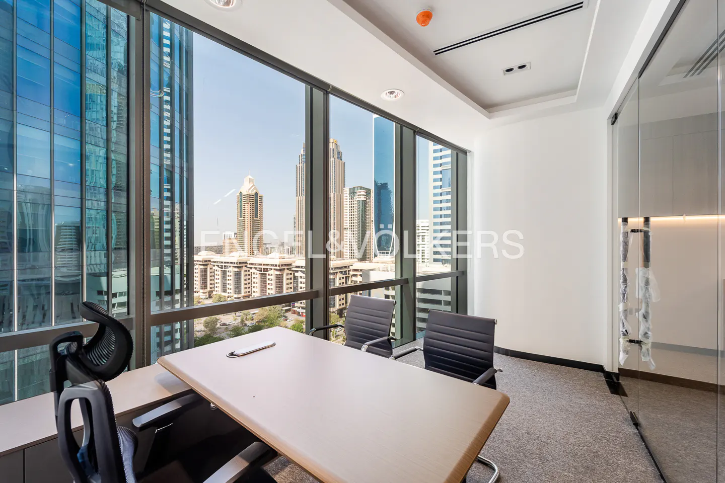 Bright office space with a large window view of city buildings. A table and chairs are in the foreground.