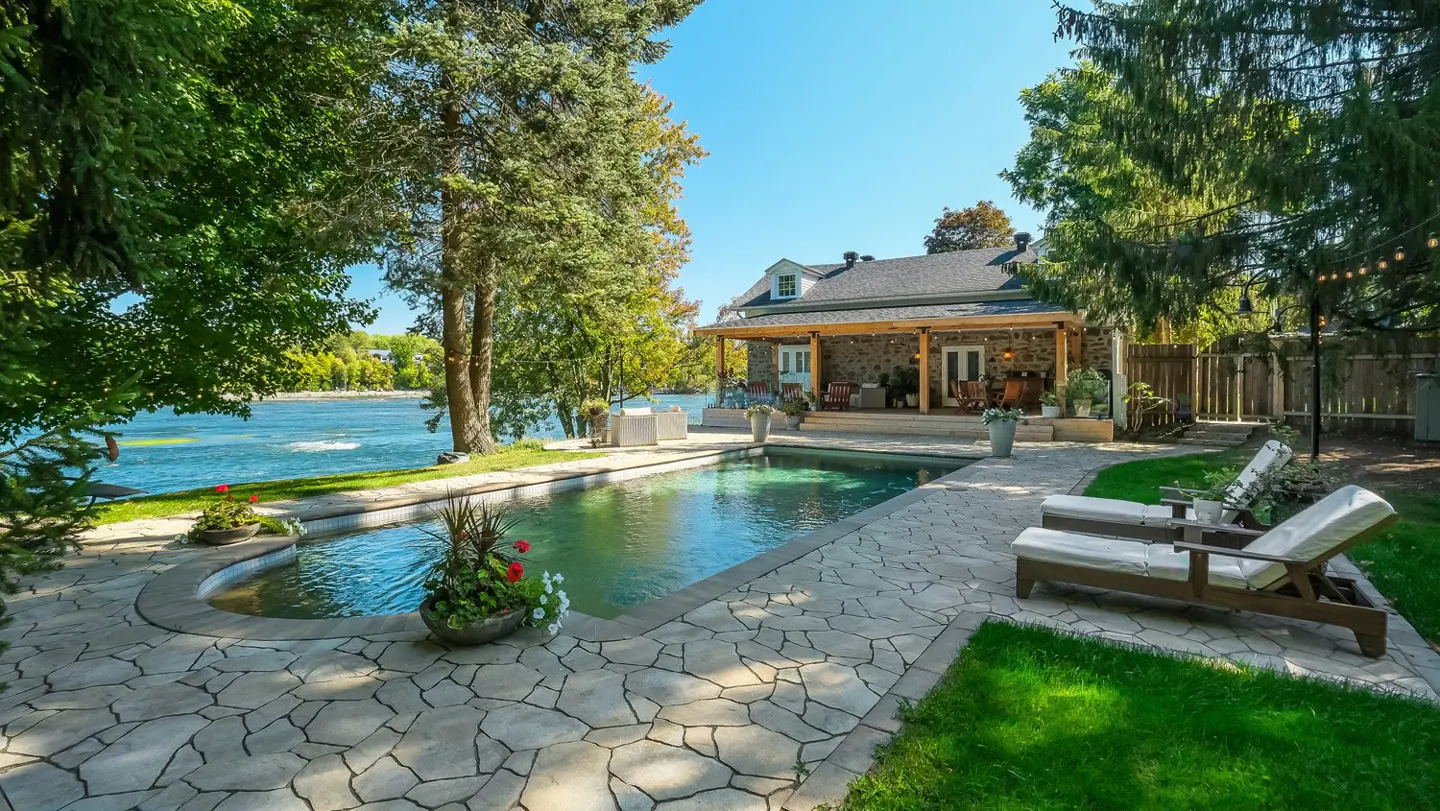 A backyard with a pool, stone patio, and lounge chairs, next to a river and a stone house with a covered porch.