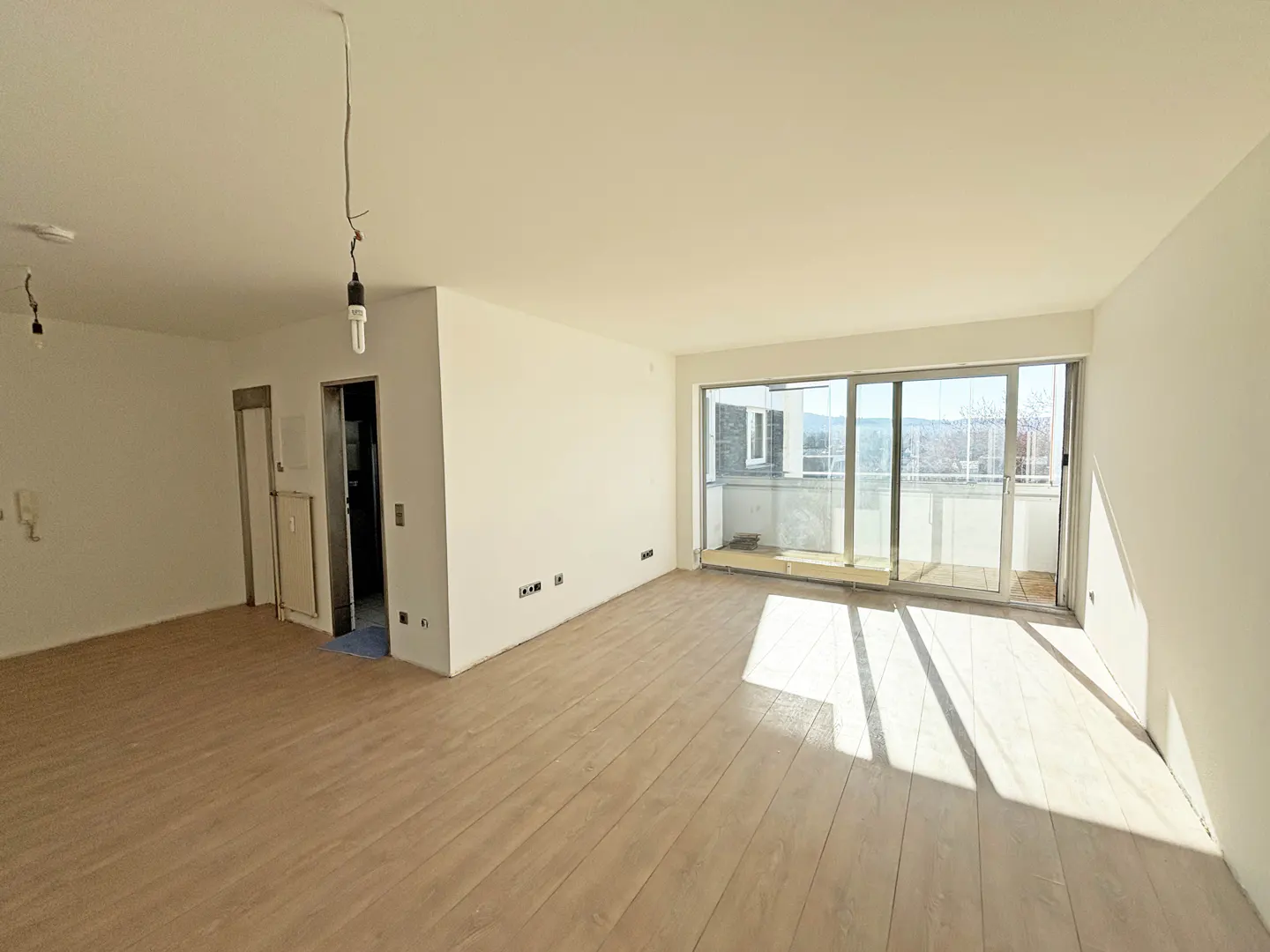 Bright, empty room with light wood floors, white walls, and a sliding glass door to a balcony. Two bare lightbulbs hang from the ceiling.