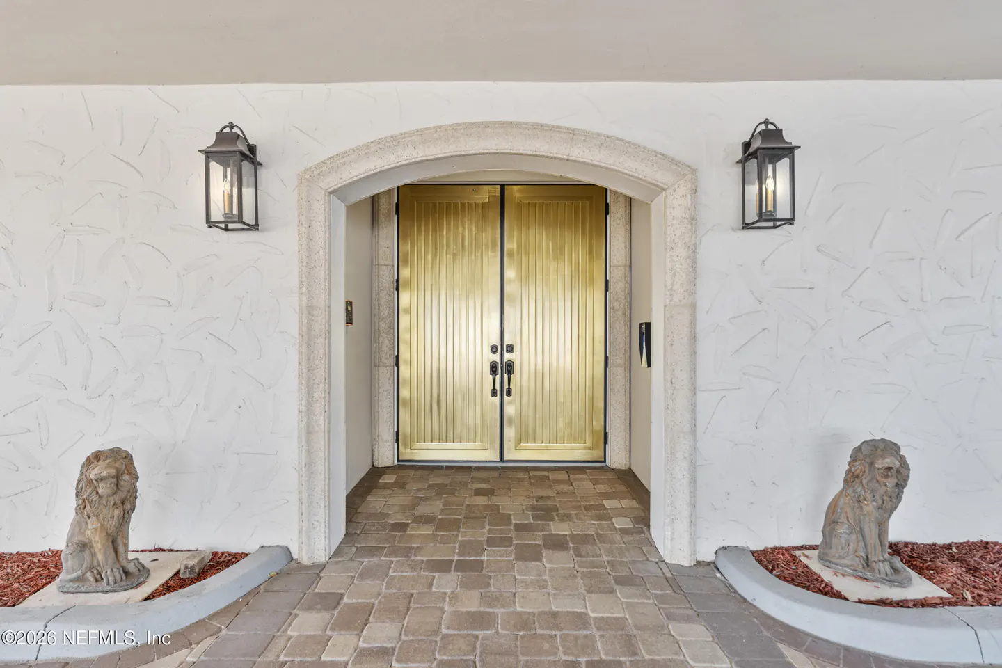Exterior view of golden double doors with stone archway, flanked by sconces and lion statues on a brick walkway.