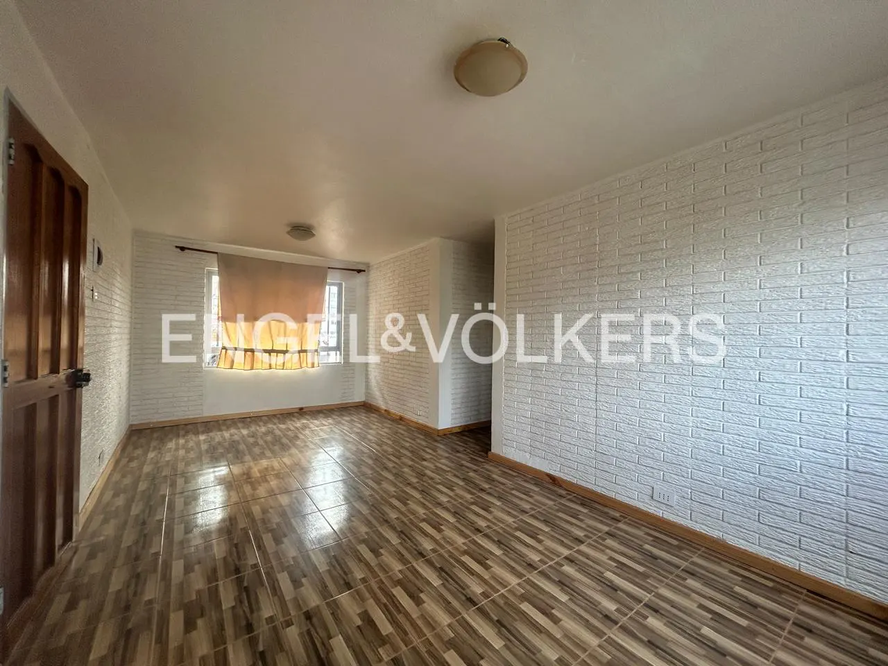 Empty room with wood-look tile floor, white brick accent wall, and a wood door on the left. Window with tan curtains. Engel & Völkers logo.