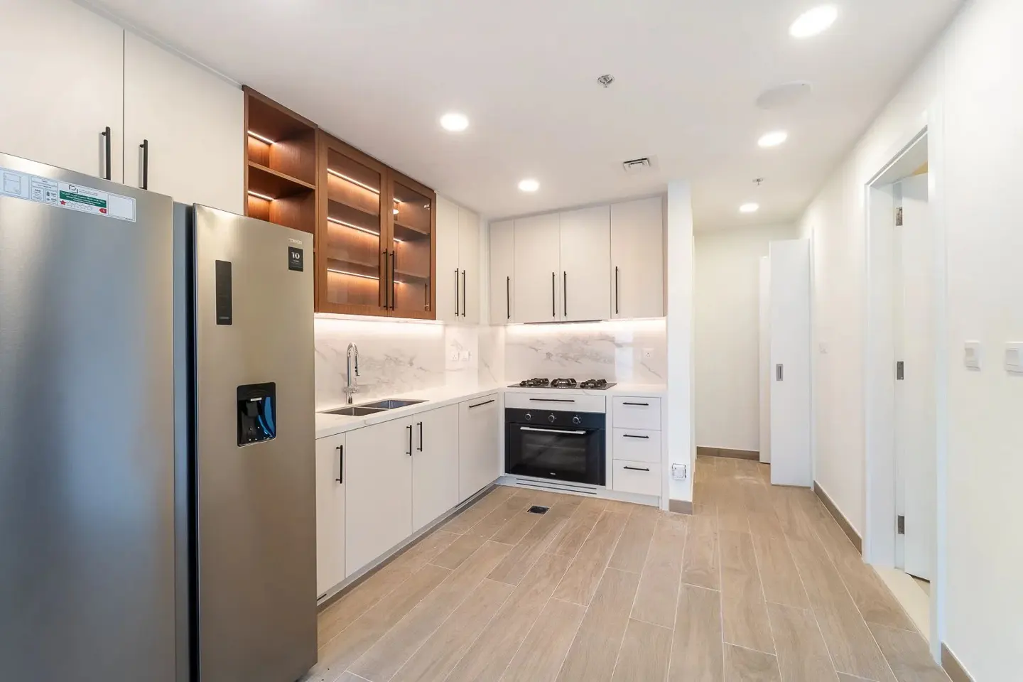 A modern kitchen with white cabinets, marble countertops, and stainless steel appliances. A hallway is visible in the background.