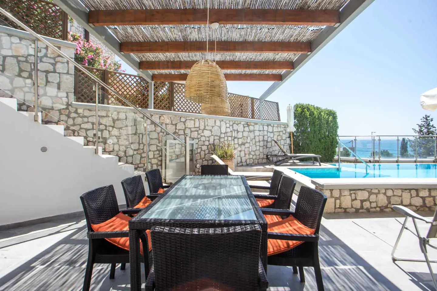 Outdoor dining area with a glass table, black wicker chairs with orange cushions, and a pool in the background.