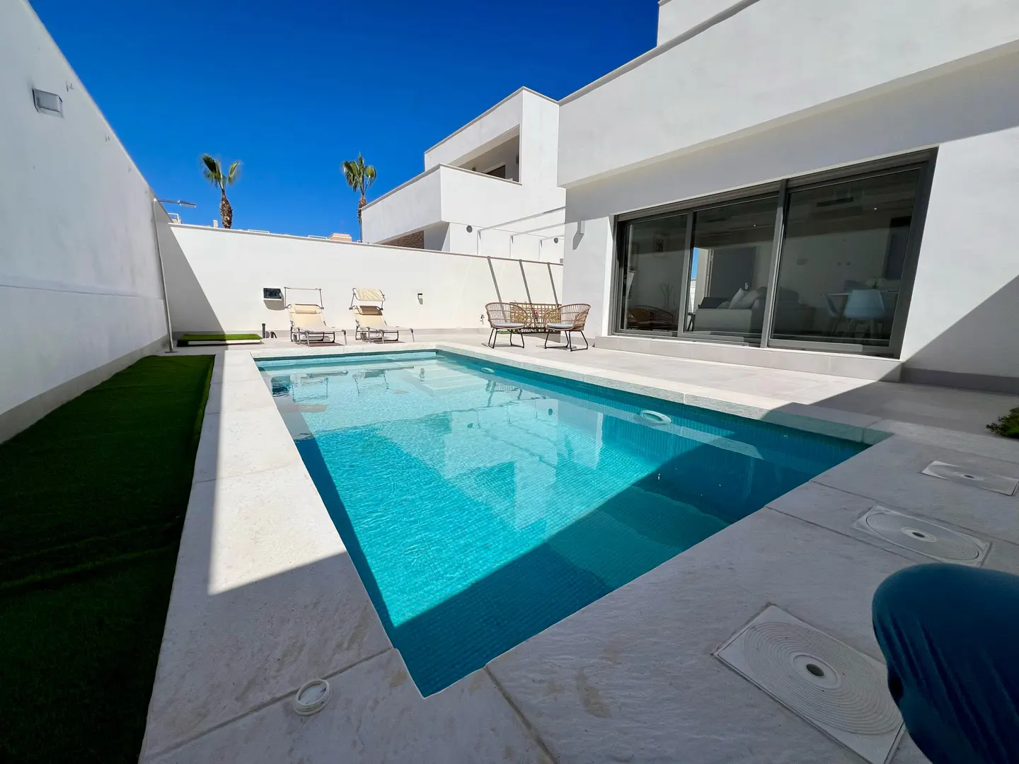 A bright blue pool is surrounded by white walls and a modern white house under a clear blue sky. Lounge chairs and a sofa are on the patio.