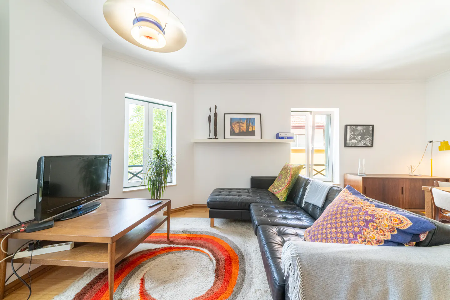 Living room with black leather sofa, wood table with TV, and orange swirl rug. White walls, windows, and modern light fixture.