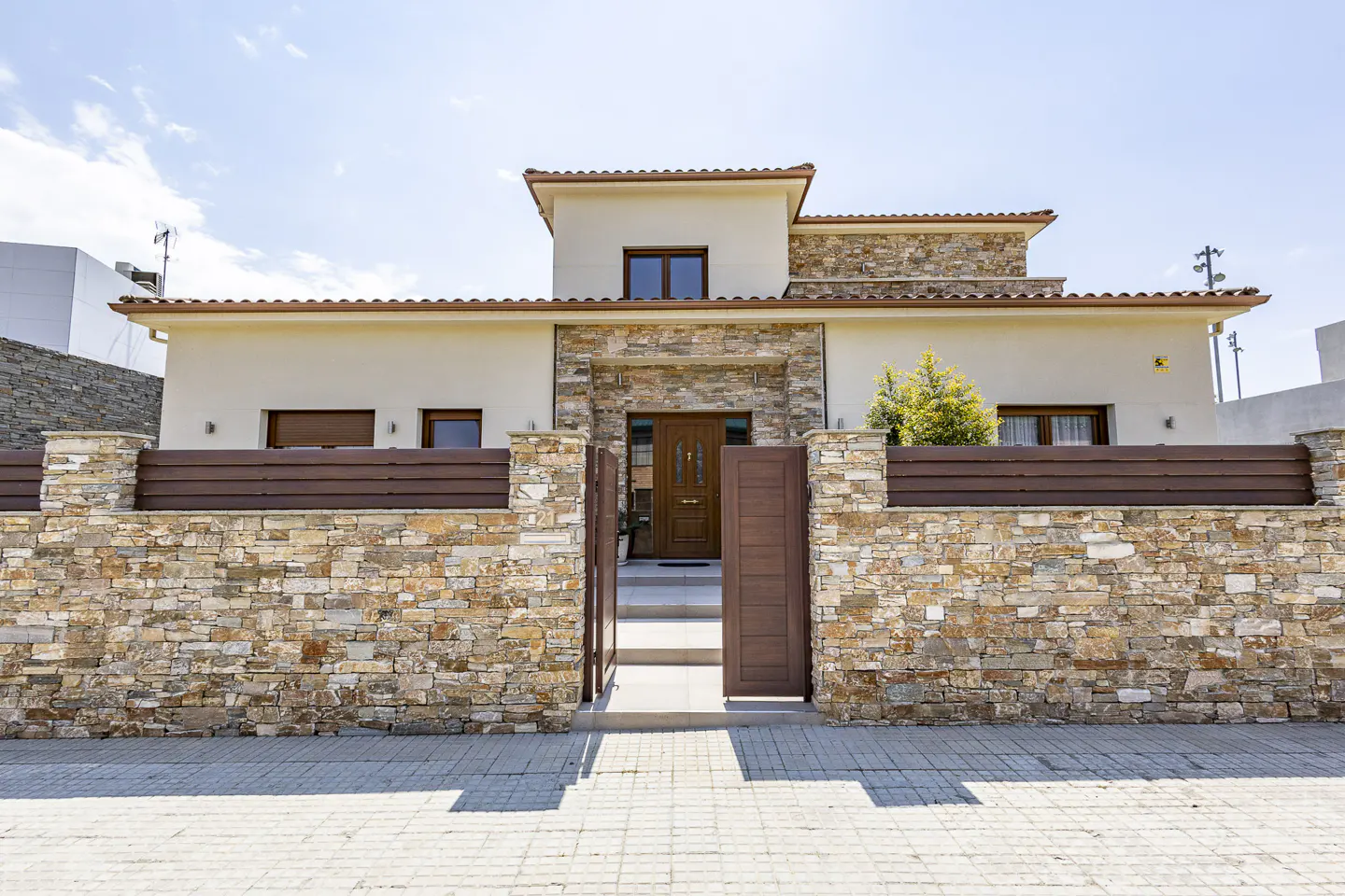 A modern, two-story house with a stone and wood fence and an open gate. The house is beige with a brown roof and stone accents.