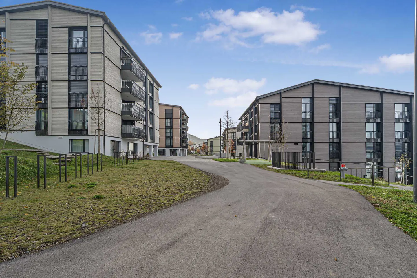 Modern apartment buildings with gray siding flank a curved asphalt road under a cloudy blue sky.