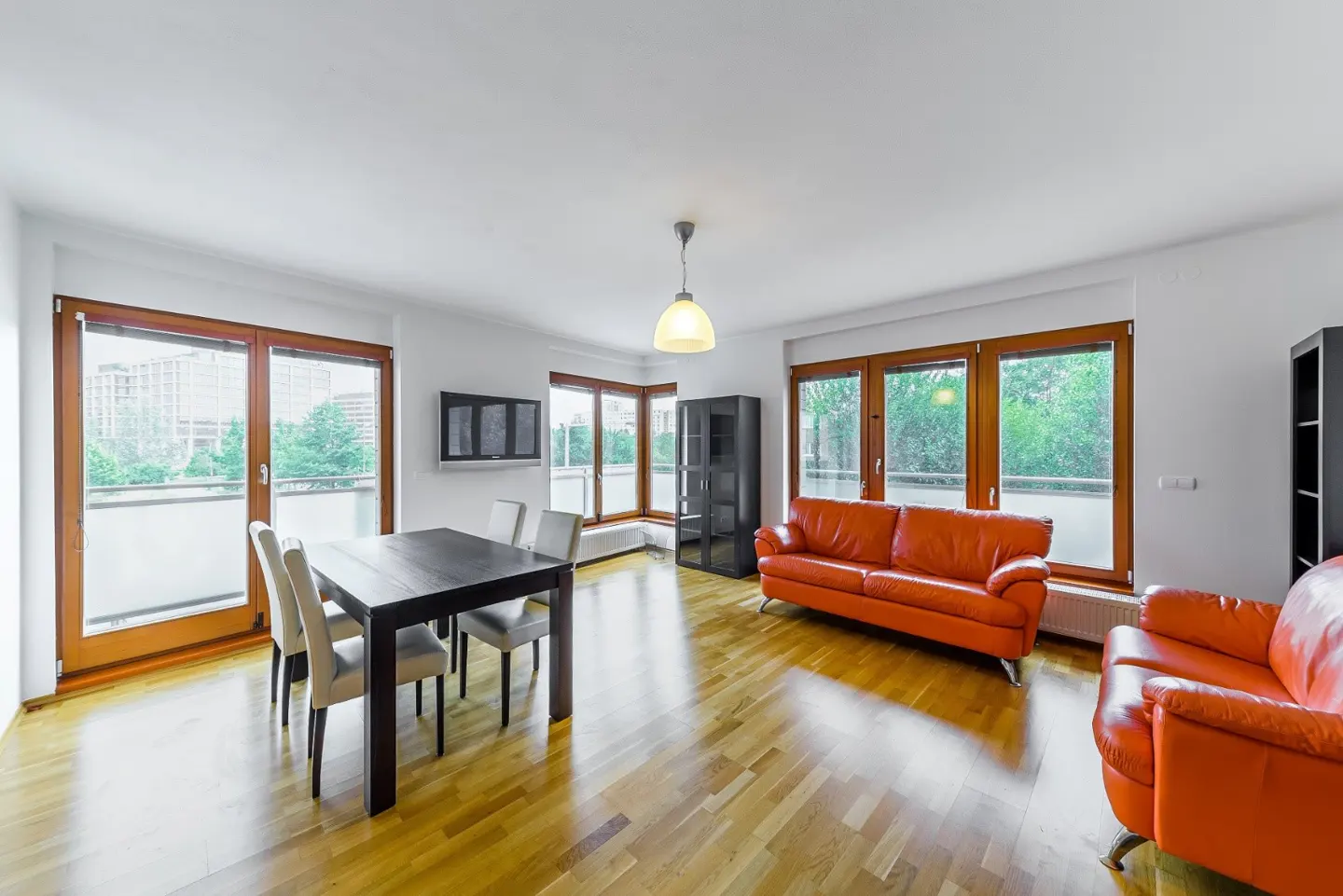 Bright living room with wood floors, orange leather sofas, a dark wood dining table with white chairs, and large windows.