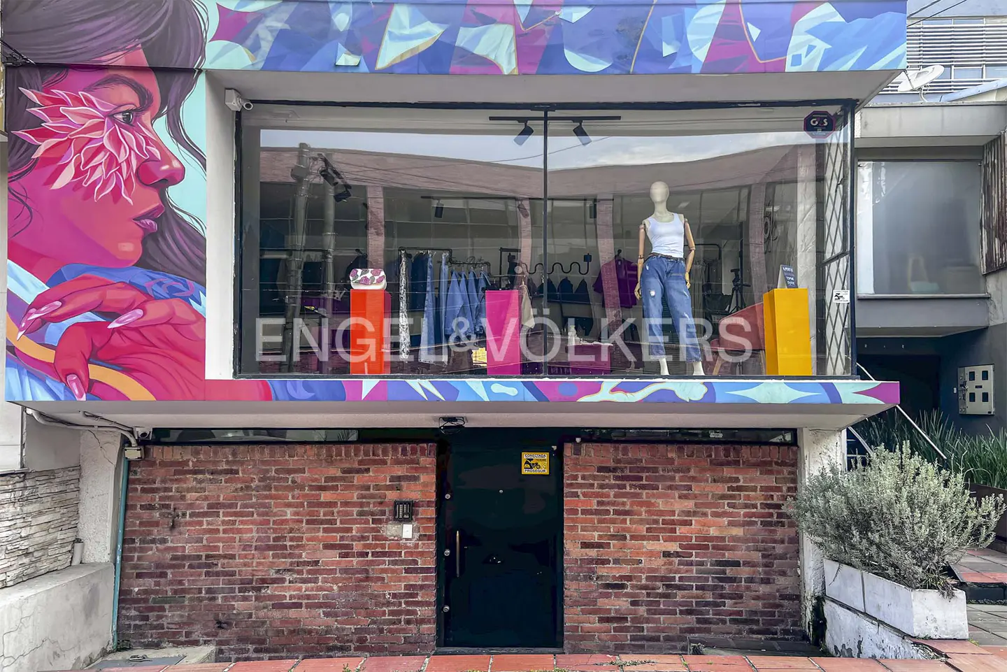 Engel & Volkers storefront with a colorful mural of a woman's face and a mannequin in the window. The building has a brick facade.
