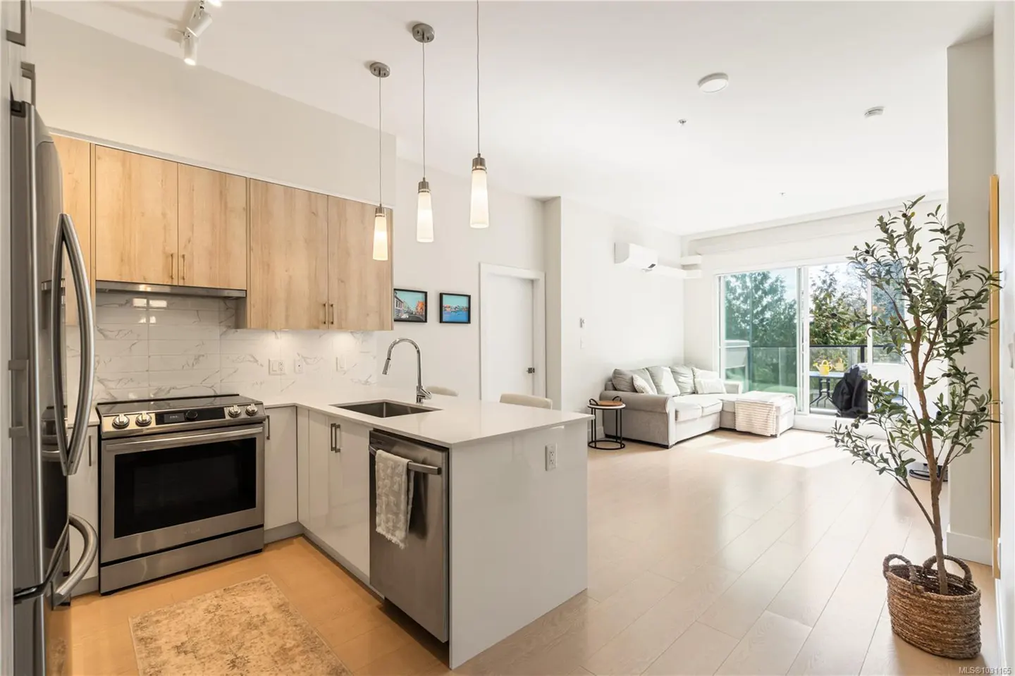Bright, open-concept condo with a kitchen featuring stainless steel appliances, light wood cabinets, and a white countertop island. A gray sofa is in the living area.