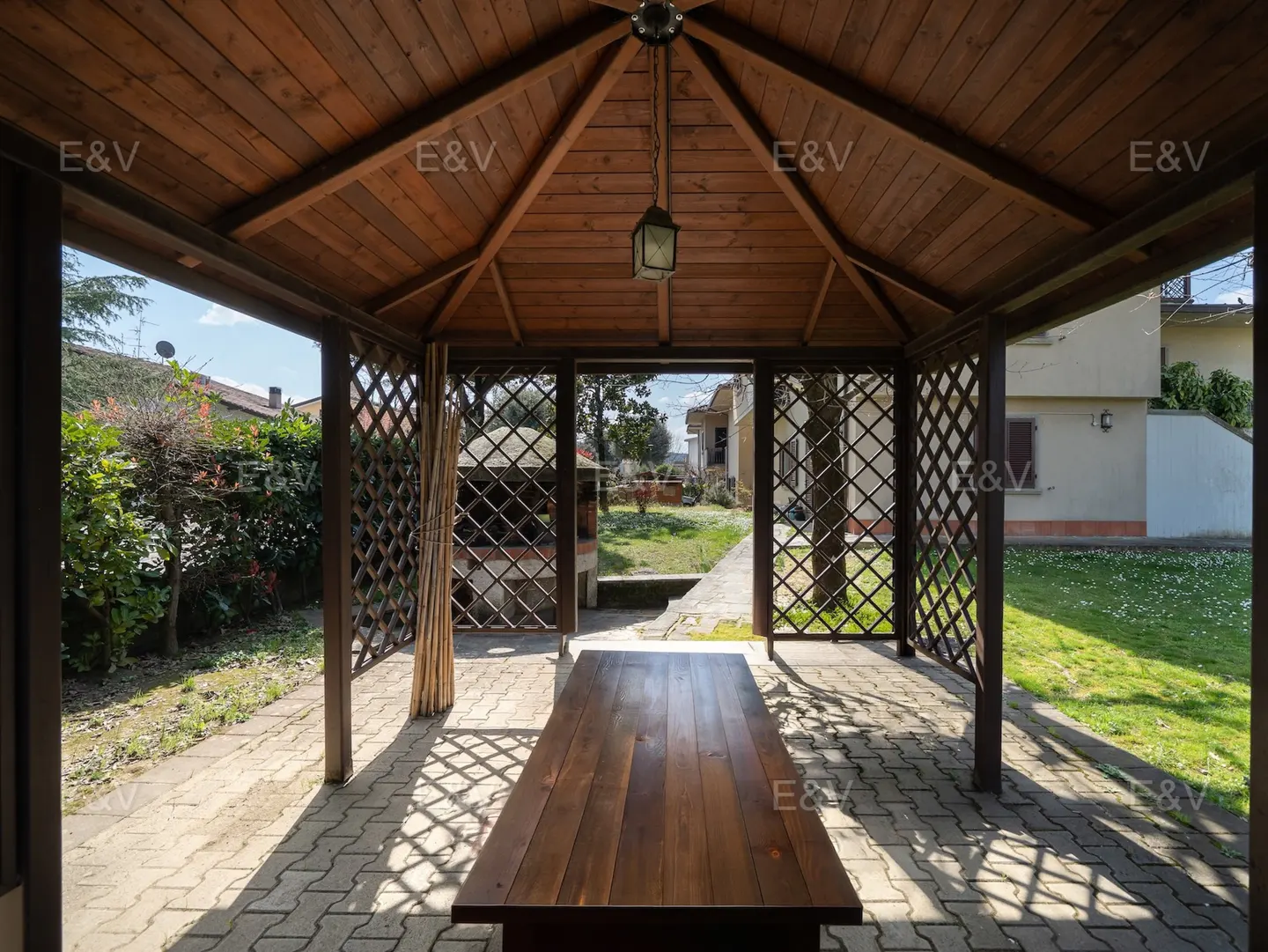 Wooden gazebo with a table inside, lattice walls, and a hanging lantern. Outside, there's a green lawn and a house.