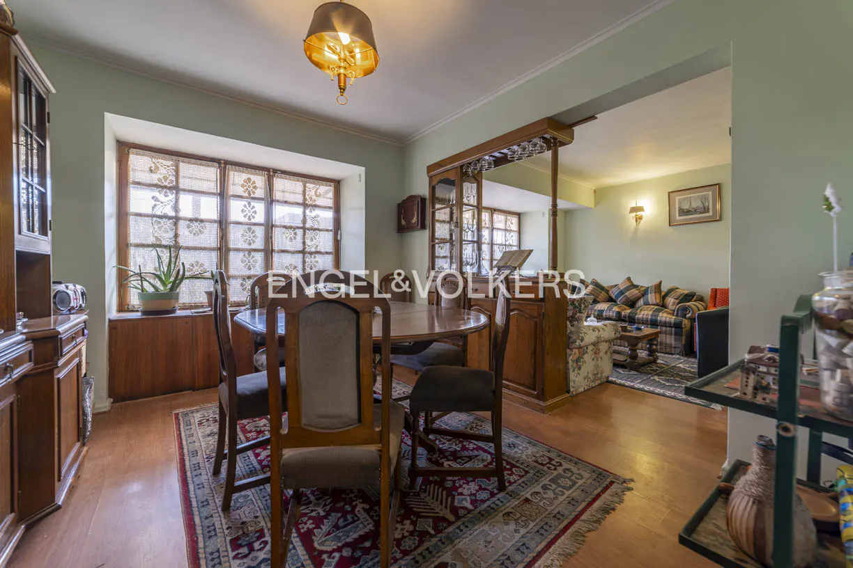 A dining room with a wooden table, chairs, and a rug, leading to a living room with a sofa.