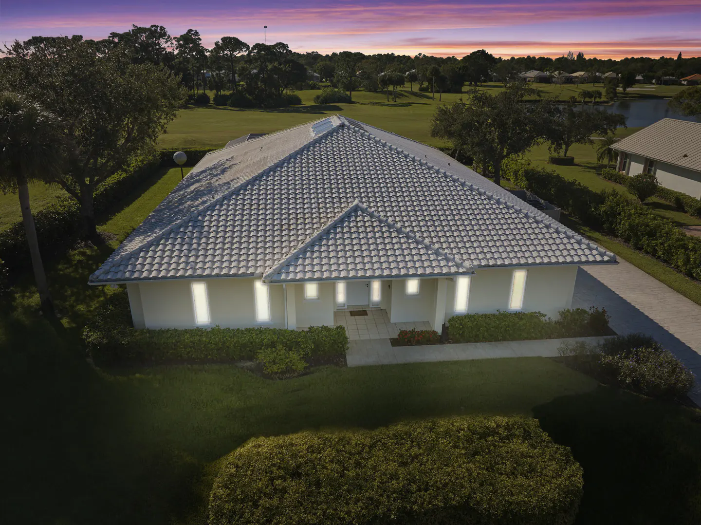 Aerial view of a one-story white house with a gray tile roof, green lawn, and trees against a pink and purple sunset sky.