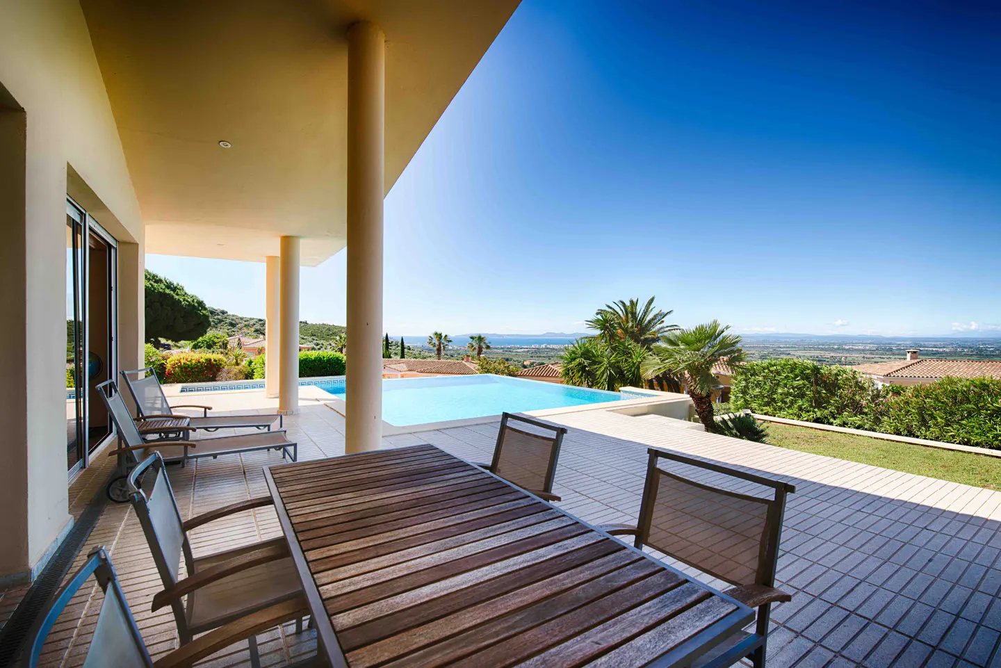 Outdoor patio with a wood table, chairs, and lounge chairs overlooking a pool, palm trees, and a distant view of the ocean under a clear blue sky.