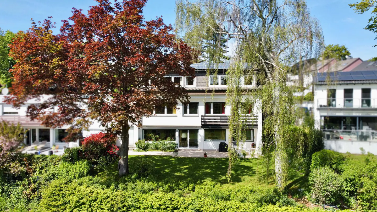 Three-story white house with a balcony, surrounded by green trees and a red-leaved tree.