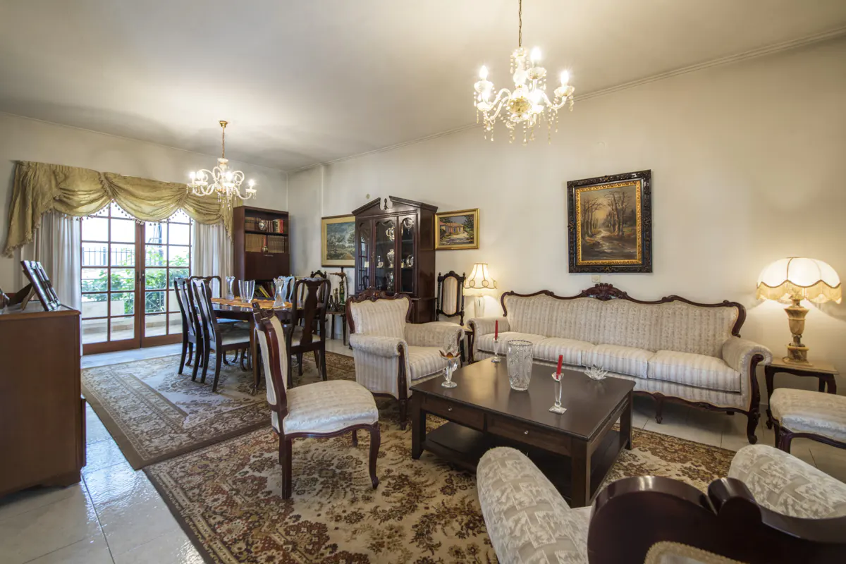 A traditionally styled living and dining room with a floral rug, chandeliers, and dark wood furniture.