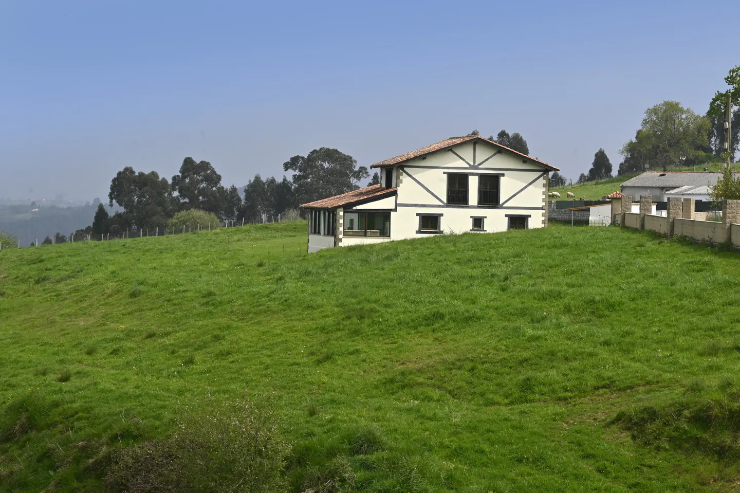 A two-story white house with black trim sits on a green hillside under a blue sky.