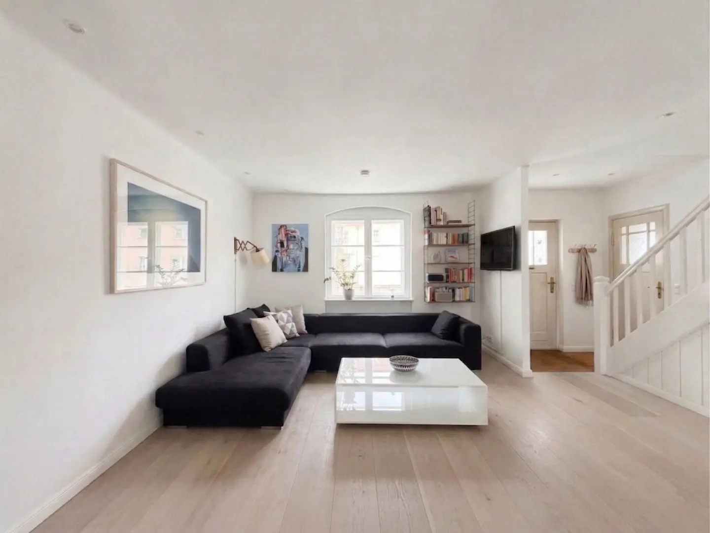 Bright living room with light wood floors, white walls, and a large black sectional sofa. A white coffee table sits in front of the sofa. Stairs are visible in the background.