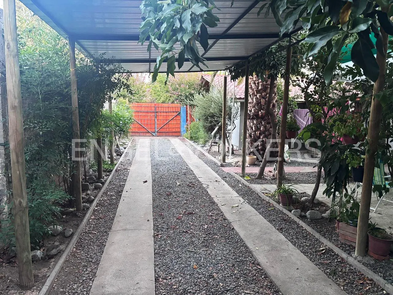 Gravel driveway leading to an orange gate, under a metal awning with trees on either side.