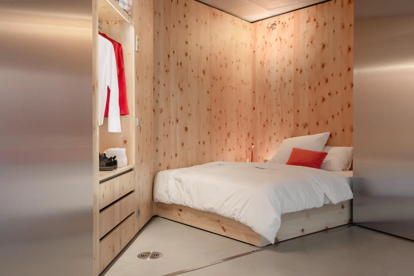 A small bedroom with wood paneled walls, a white bed with an orange pillow, and an open closet with clothes.