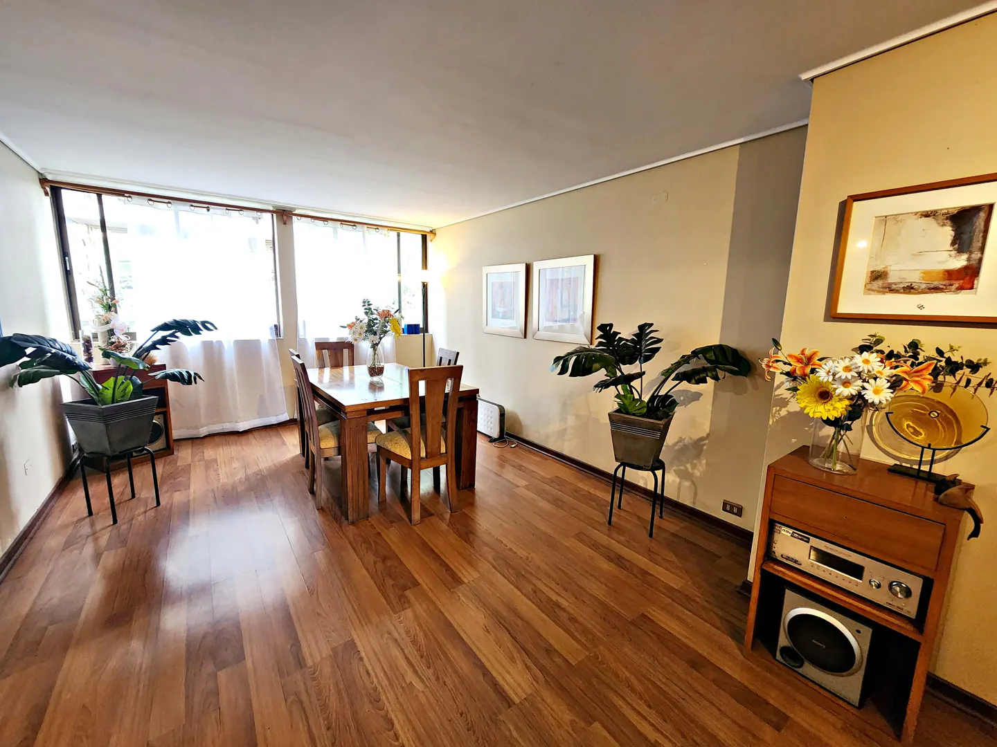 A dining room with a wooden table and chairs, hardwood floors, and beige walls. There are plants and artwork throughout the room.