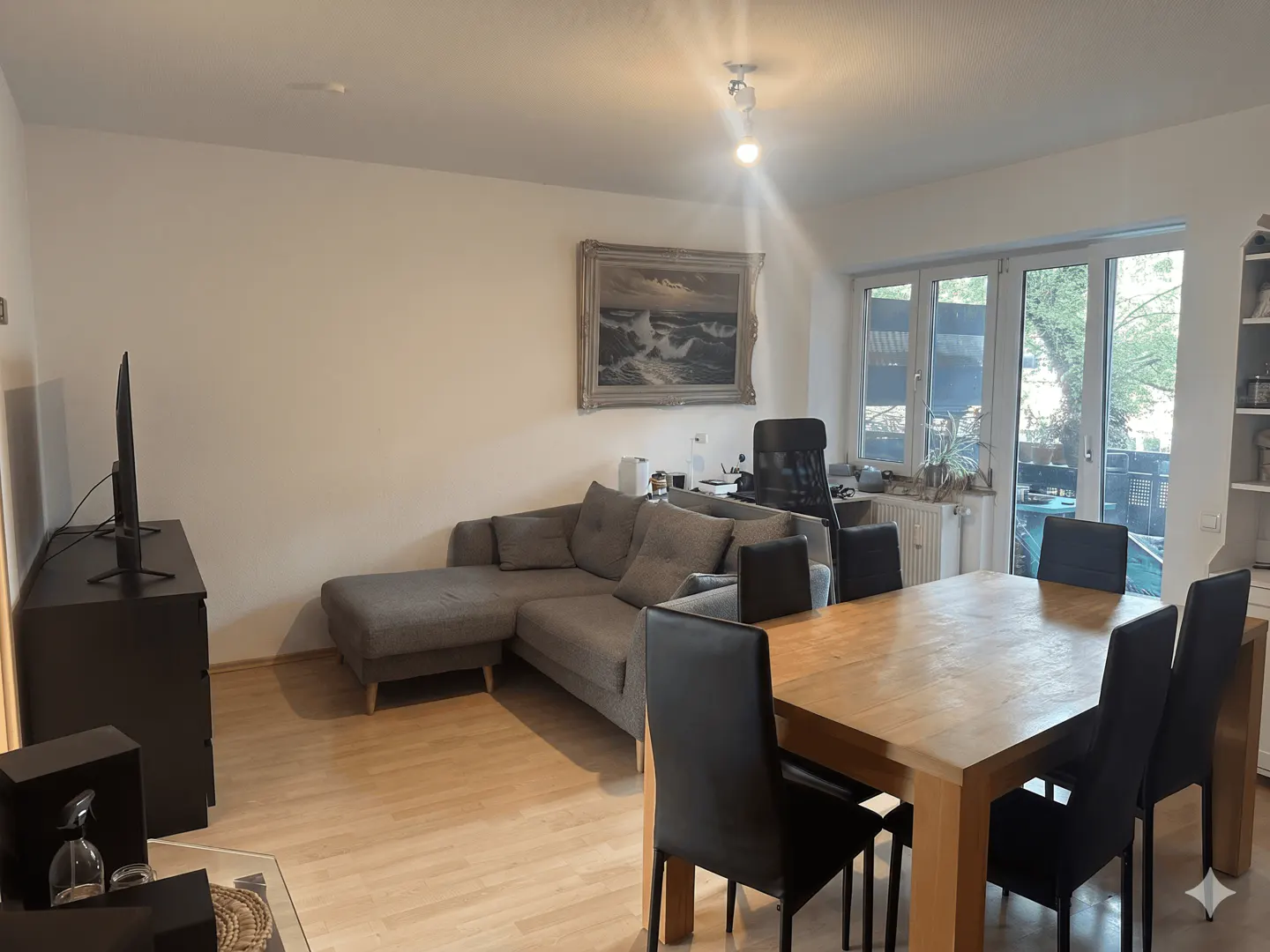 Living room with a grey sectional sofa, wooden dining table with black chairs, and a framed seascape painting on the wall.