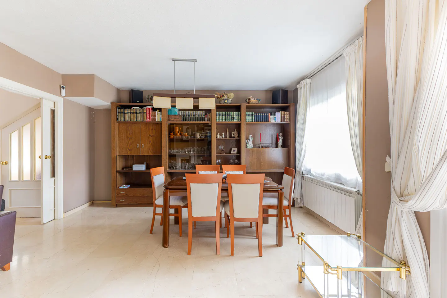 A dining room with a wooden table and chairs, a large bookcase, and a window with white curtains.