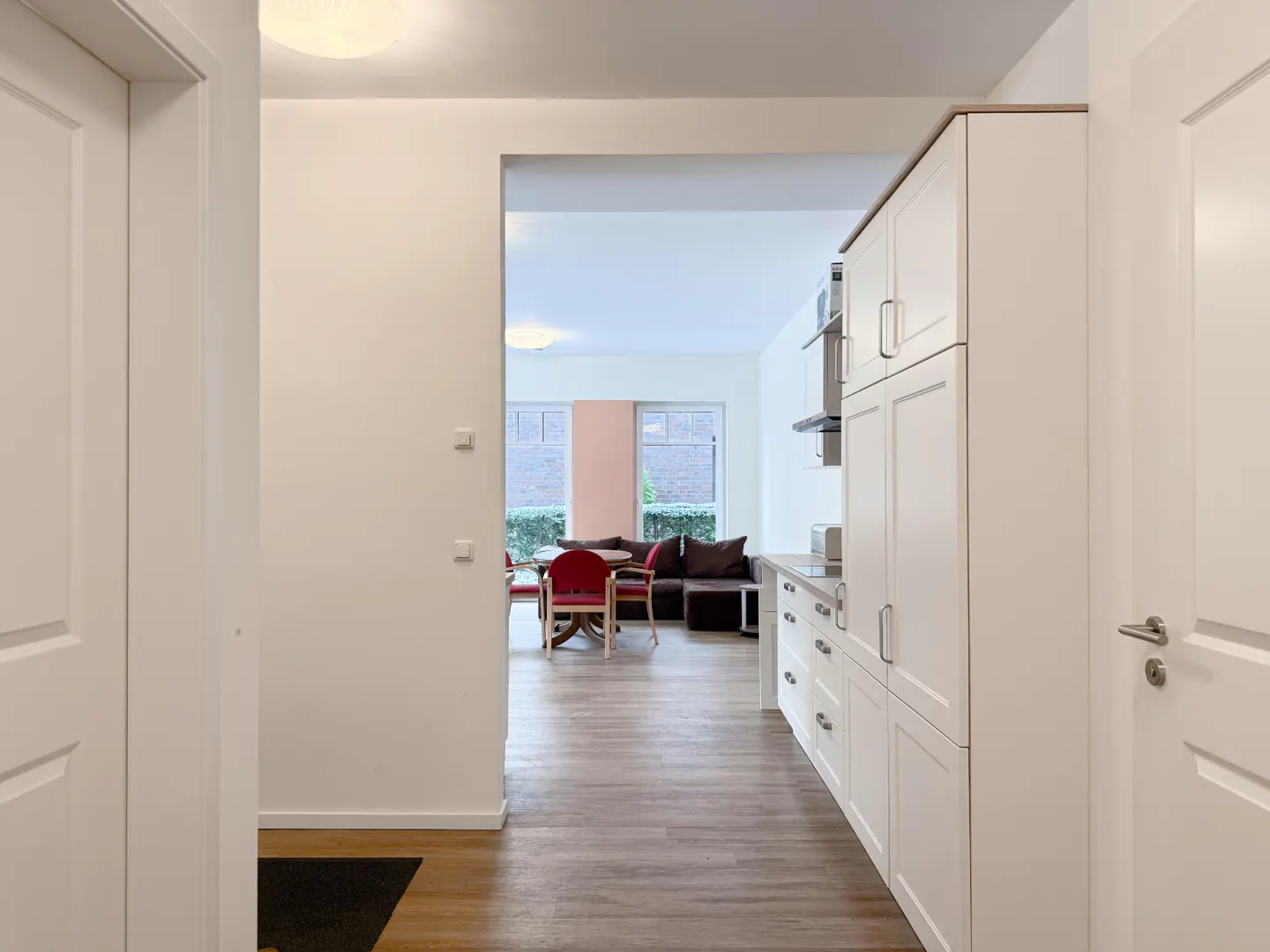 Hallway view into a bright living space with wood floors, white walls, and white kitchen cabinets. A dining table and brown sofa are visible.