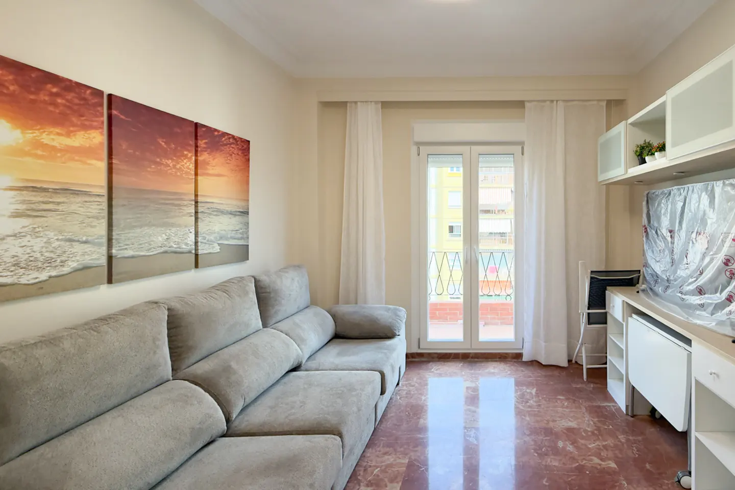 Living room with a gray sofa, a beach triptych, and a balcony with white curtains. The floor is red marble.