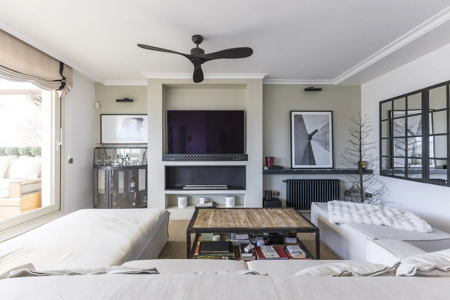 Living room with white sofas, a wood and metal coffee table, and a built-in entertainment center with a TV and shelves. A black ceiling fan hangs above.