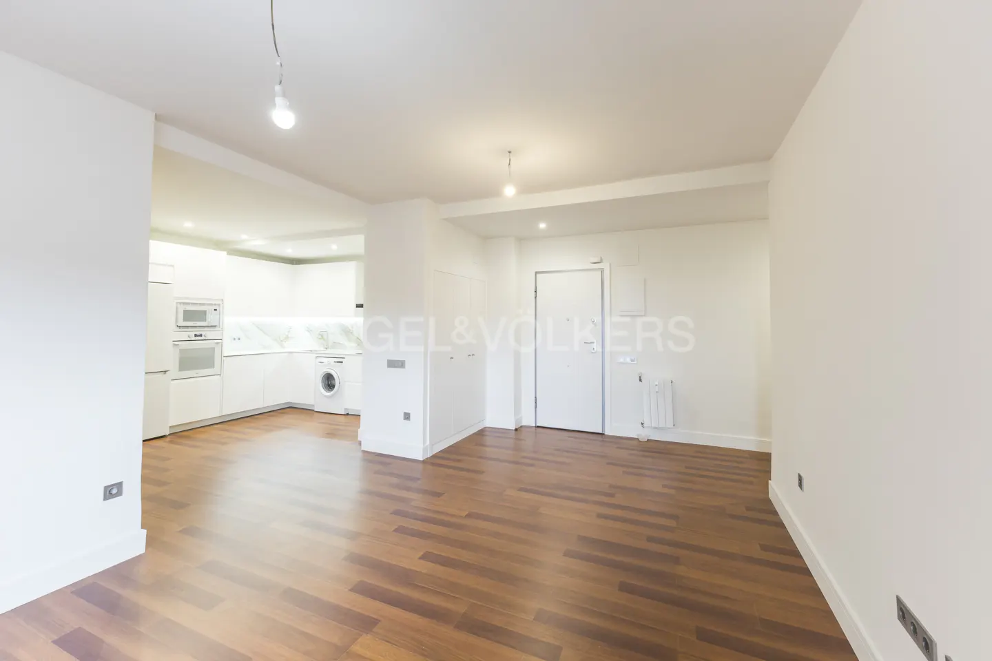 Bright, open-concept apartment with hardwood floors and white walls. Kitchen with white appliances and cabinets is visible in the background.