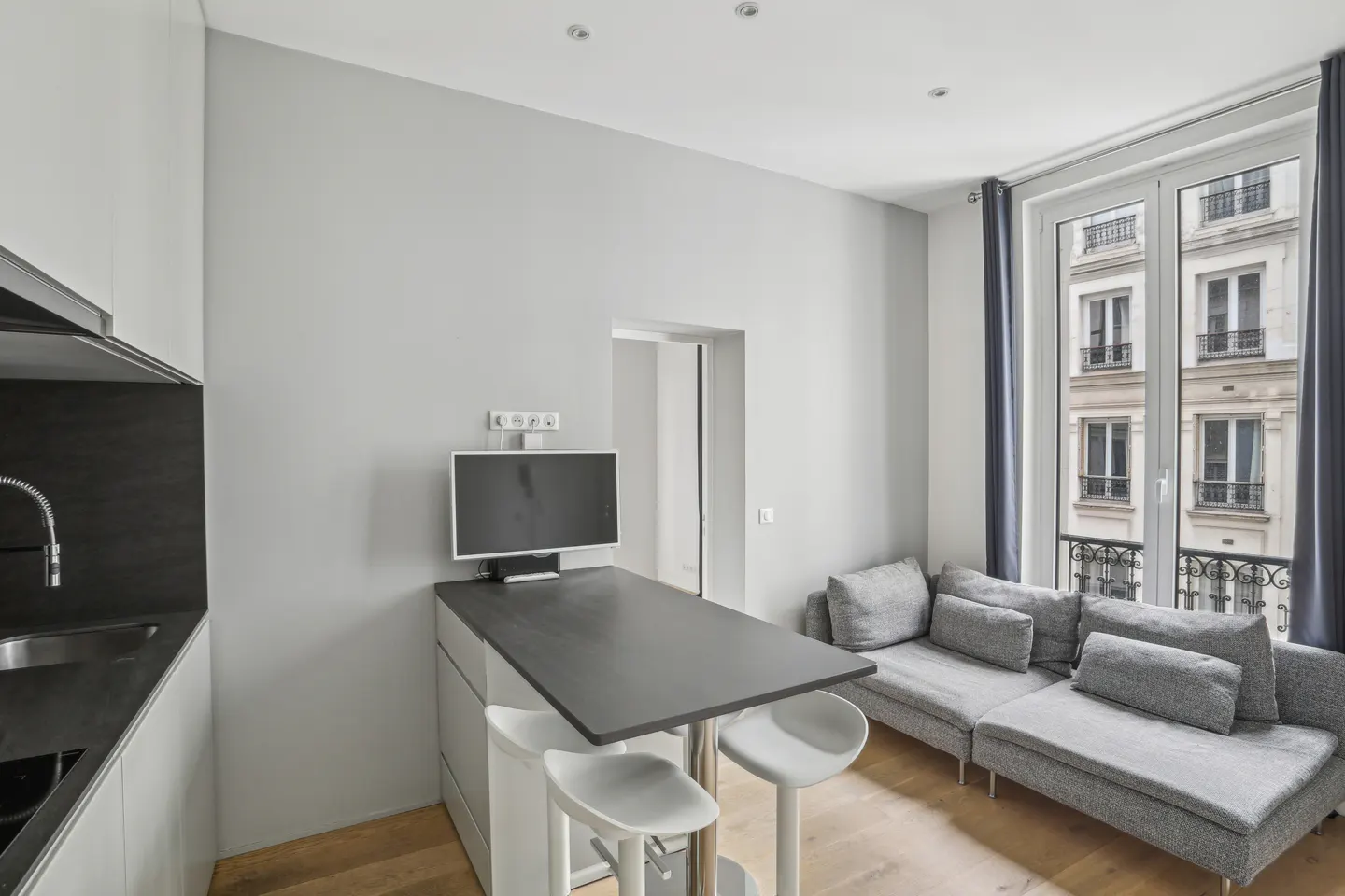 A modern apartment interior with a gray sofa, a black countertop with white stools, and a window view of a building.